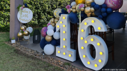 Large illuminated numbers "16" are displayed next to a cluster of balloons in various colors, including gold, white, and blue. In the background, a sign reads "Feliz cumpleaños.