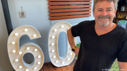 Man smiling next to large, illuminated "60" sign at an outdoor gathering.